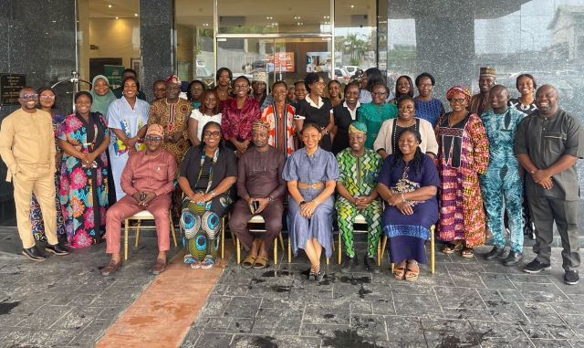 Oyo PHC Large diverse group of adults in colorful traditional clothing posing for a group photo outside a glass-front building; some seated in front and others standing behind them.