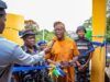 We’ll Continue To Provide Necessary Support To Security Agencies—Makinde Man in orange traditional attire speaks into a microphone while cutting a blue ribbon at a ceremony, flanked by police officers and onlookers near a yellow structure.