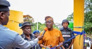We’ll Continue To Provide Necessary Support To Security Agencies—Makinde Man in orange traditional attire speaks into a microphone while cutting a blue ribbon at a ceremony, flanked by police officers and onlookers near a yellow structure.