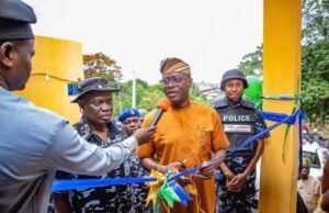 We’ll Continue To Provide Necessary Support To Security Agencies—Makinde Man in orange traditional attire speaks into a microphone while cutting a blue ribbon at a ceremony, flanked by police officers and onlookers near a yellow structure.
