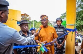 We’ll Continue To Provide Necessary Support To Security Agencies—Makinde Man in orange traditional attire speaks into a microphone while cutting a blue ribbon at a ceremony, flanked by police officers and onlookers near a yellow structure.