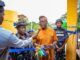 We’ll Continue To Provide Necessary Support To Security Agencies—Makinde Man in orange traditional attire speaks into a microphone while cutting a blue ribbon at a ceremony, flanked by police officers and onlookers near a yellow structure.