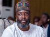 Tinubu Appoints Prof Segun Aina As New JAMB Registrar Man with gray beard wearing a colorful patterned cap and white traditional tunic, seated indoors at an event.