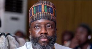 Tinubu Appoints Prof Segun Aina As New JAMB Registrar Man with gray beard wearing a colorful patterned cap and white traditional tunic, seated indoors at an event.