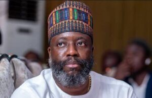 Tinubu Appoints Prof Segun Aina As New JAMB Registrar Man with gray beard wearing a colorful patterned cap and white traditional tunic, seated indoors at an event.