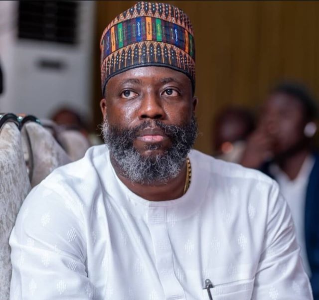 Professor Segun Aina Man with gray beard wearing a colorful patterned cap and white traditional tunic, seated indoors at an event.