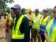 RAAMP: Oyo Govt Completes 88.02 Km Of Rural Roads, Markets In Phase One Of Project Group of people in high-visibility vests at a construction site, a man in a cap speaks into a microphone while others listen.