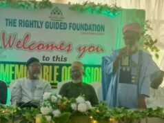 UI Imam Tasks Muslims With Kindness, Good Deeds To Humanity Man in blue traditional attire speaks at a foundation event on a decorated stage; banner behind reads 'The Rightly Guided Foundation welcomes you to this memorable session'.