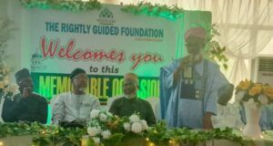 UI Imam Tasks Muslims With Kindness, Good Deeds To Humanity Man in blue traditional attire speaks at a foundation event on a decorated stage; banner behind reads 'The Rightly Guided Foundation welcomes you to this memorable session'.