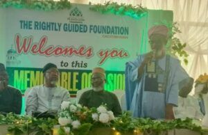 UI Imam Tasks Muslims With Kindness, Good Deeds To Humanity Man in blue traditional attire speaks at a foundation event on a decorated stage; banner behind reads 'The Rightly Guided Foundation welcomes you to this memorable session'.