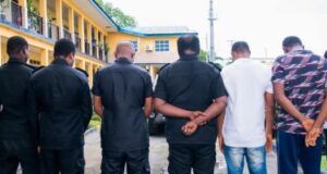 Rivers Police Dismiss, Prosecute Five Officers Over Alleged Armed Robbery, Kidnapping Group of men standing with their backs to the camera, arms folded behind their backs, in a courtyard outside a yellow building with balconies.