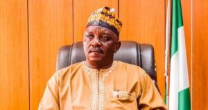 Court Sentences Ex-Power Minister Mamman To 75 Years Man wearing traditional beige attire and a patterned cap sits at a desk in a formal office, with a Nigerian flag in the background.