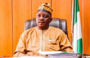 Court Sentences Ex-Power Minister Mamman To 75 Years Man wearing traditional beige attire and a patterned cap sits at a desk in a formal office, with a Nigerian flag in the background.