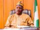 Court Sentences Ex-Power Minister Mamman To 75 Years Man wearing traditional beige attire and a patterned cap sits at a desk in a formal office, with a Nigerian flag in the background.