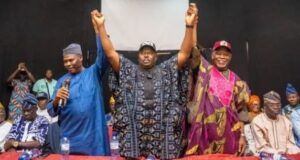 APC Leaders Endorse Senator Alli As Consensus Governorship Candidate For Oyo 2027 Three men on a stage raise their arms in victory, flanked by supporters in traditional clothing.