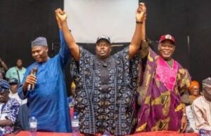 APC Leaders Endorse Senator Alli As Consensus Governorship Candidate For Oyo 2027 Three men on a stage raise their arms in victory, flanked by supporters in traditional clothing.