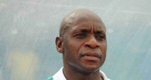 Reasons Abia Warriors Collapsed In 2025/2026 NPFL Top Three Race—Shorunmu Man with a shaved head wearing a white polo with green stripes, looking to the side outdoors against a blue background.