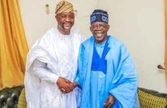 Tinubu Took Tough Decisions Past Leaders Avoided — Sunday Dare Two men wearing white and blue agbadas shake hands and smile in a living room setting.