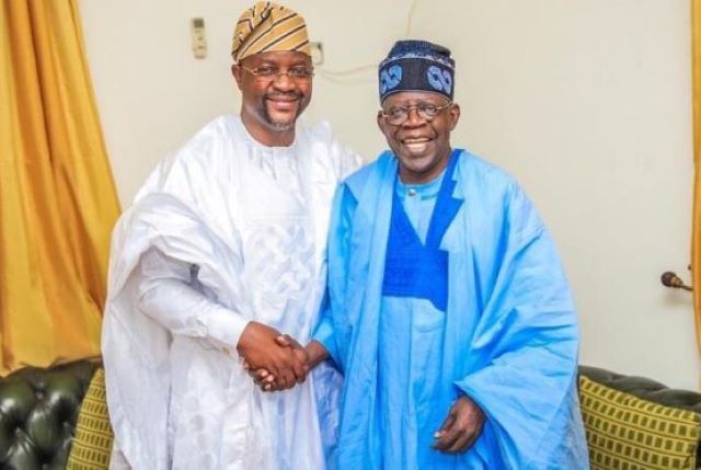 Sunday Dare with President Tinubu Two men wearing white and blue agbadas shake hands and smile in a living room setting.