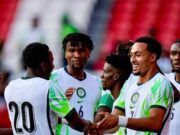 Unity Cup: Lawal Gives Thumbs Up To Eagles’ Victory Against Zimbabwe Soccer players in white jerseys with green accents celebrate, shaking hands on the field with red stadium seats in the background.