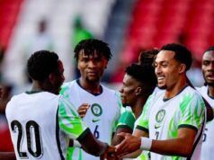 Unity Cup: Lawal Gives Thumbs Up To Eagles’ Victory Against Zimbabwe Soccer players in white jerseys with green accents celebrate, shaking hands on the field with red stadium seats in the background.