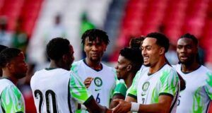 Unity Cup: Lawal Gives Thumbs Up To Eagles’ Victory Against Zimbabwe Soccer players in white jerseys with green accents celebrate, shaking hands on the field with red stadium seats in the background.