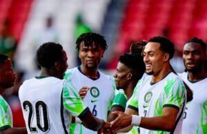 Unity Cup: Lawal Gives Thumbs Up To Eagles’ Victory Against Zimbabwe Soccer players in white jerseys with green accents celebrate, shaking hands on the field with red stadium seats in the background.