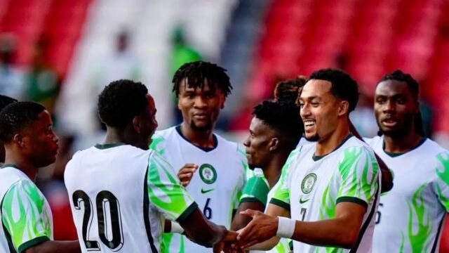 Soccer players in white jerseys with green accents celebrate, shaking hands on the field with red stadium seats in the background.