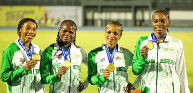 Team Nigeria Relay Four female athletes in green-and-white jackets hold gold medals, smiling on a track field during an awards ceremony.