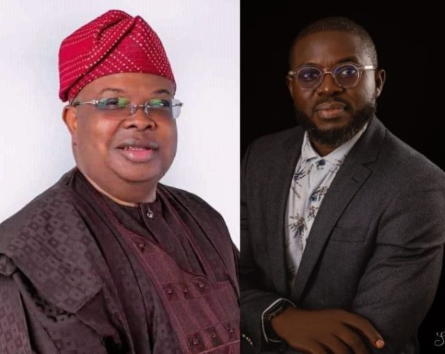 Tegbe and Giwa Two men seated side by side; left in traditional maroon attire with a red patterned cap, right in a gray suit and glasses.