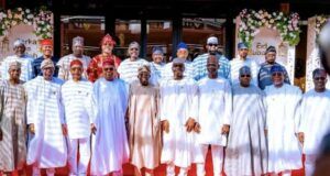 I’m Happy That There Is No State Governor That Struggles To Pay Salaries—Tinubu Group of men in traditional Nigerian attire posing for a photo on a red carpet at an Eid celebration, with floral decorations in the background.