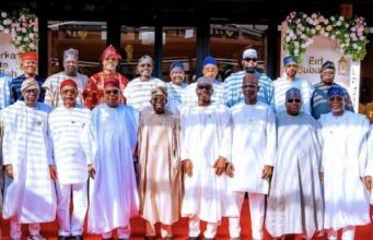 I’m Happy That There Is No State Governor That Struggles To Pay Salaries—Tinubu Group of men in traditional Nigerian attire posing for a photo on a red carpet at an Eid celebration, with floral decorations in the background.