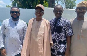 Four men standing side by side outdoors smiling at the camera they wear a mix of traditional and casual clothing with a white wall and palm trees behind them
