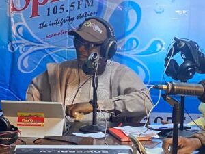 Radio host wearing a cap and headphones speaks into a microphone at a broadcasting desk with a blue backdrop showing 1055FM