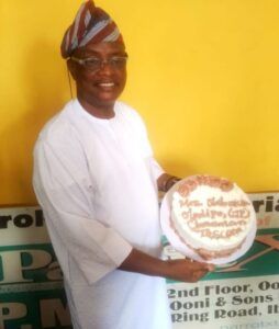 Man in a white traditional outfit and striped head wrap smiles while holding a frosted cake with brown icing against a yellow wall