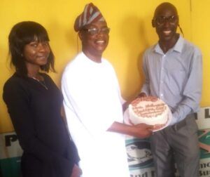 Three adults pose with a frosted round cake in front of a yellow wall center person in white traditional attire holds the cake flanked by a woman on the left and a man on the right all smiling