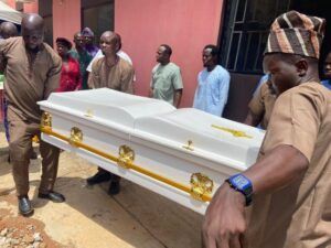 Casket being carried by pallbearers in brown outfits, with onlookers beside a pink wall at a funeral site.