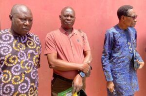 Three men stand against a pink wall; left in a purple-yellow patterned shirt, center in a red polo, right in a blue patterned outfit with sunglasses.