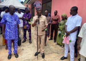 Man in a beige suit speaks into a microphone at a community event, as a crowd in colorful traditional clothing watches outside a pink building.