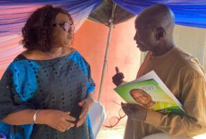 Two adults talk under a blue canopy; a woman in a blue dress wearing sunglasses listens as a man in tan holds a green pamphlet.