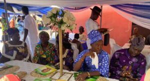 Group of people in colorful traditional attire sitting under a blue-and-white tent at a social event, with a floral centerpiece and green booklets on a table.