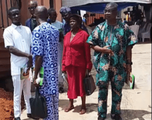 Group of people standing outdoors in front of a building, some in bright traditional African attire, center woman in red suit holding a bag/phone.