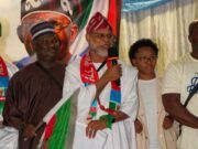 APC-South Africa Inaugurates Mpumalanga Executives, Strengthens Mobilisation For 2027 Election Man in white traditional robe speaks into a microphone at a cultural event, draped in a green, red, and white flag with others nearby.