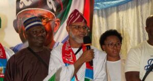 APC-South Africa Inaugurates Mpumalanga Executives, Strengthens Mobilisation For 2027 Election Man in white traditional robe speaks into a microphone at a cultural event, draped in a green, red, and white flag with others nearby.