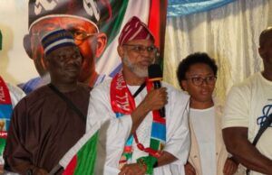 APC-South Africa Inaugurates Mpumalanga Executives, Strengthens Mobilisation For 2027 Election Man in white traditional robe speaks into a microphone at a cultural event, draped in a green, red, and white flag with others nearby.
