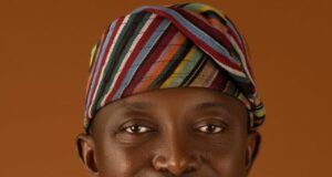 Oyo Warns Residents Of Flash Floods, Calls For Strict Adherence To Safety Measures Portrait of a smiling man wearing a colorful striped headwrap and white traditional attire against a warm brown background.