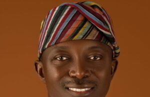 Oyo Warns Residents Of Flash Floods, Calls For Strict Adherence To Safety Measures Portrait of a smiling man wearing a colorful striped headwrap and white traditional attire against a warm brown background.