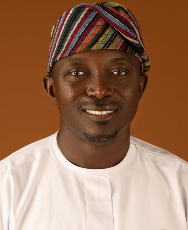 Aderinto Portrait of a smiling man wearing a colorful striped headwrap and white traditional attire against a warm brown background.
