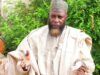 Unpaid N9m Debt: Alhaji Akewugbagold To Face Legal Action Man in beige traditional robe and patterned cap gestures with his hands outdoors, green bushes in the background.