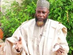 Unpaid N9m Debt: Alhaji Akewugbagold To Face Legal Action Man in beige traditional robe and patterned cap gestures with his hands outdoors, green bushes in the background.